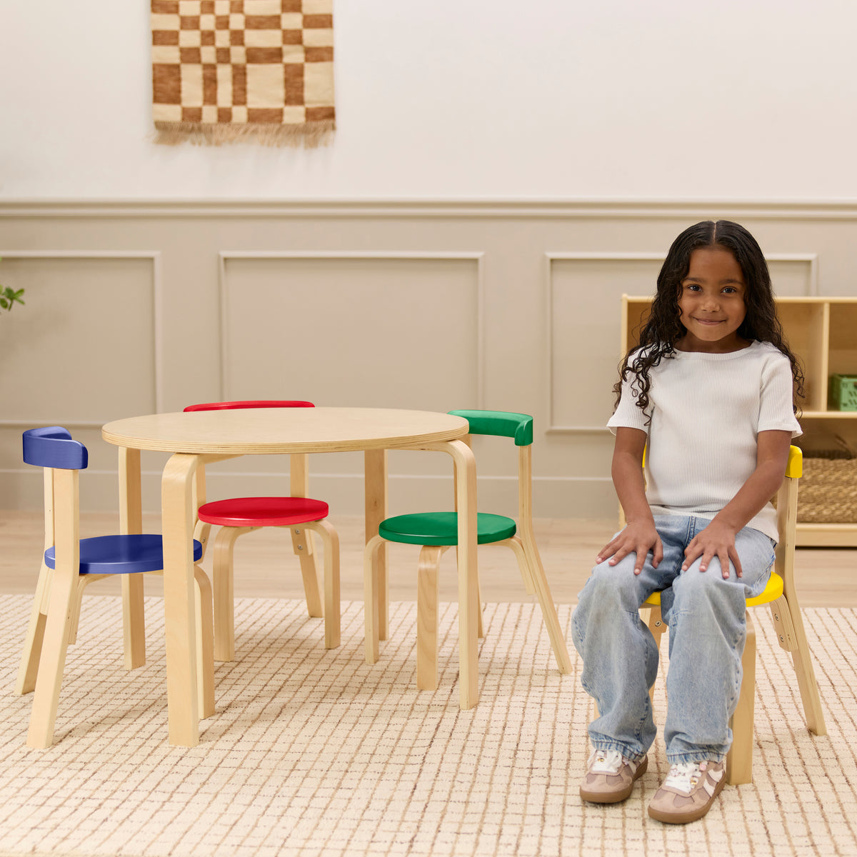 Natural wood round table with four chairs featuring blue, red, and green padded seats arranged in a playroom setting.