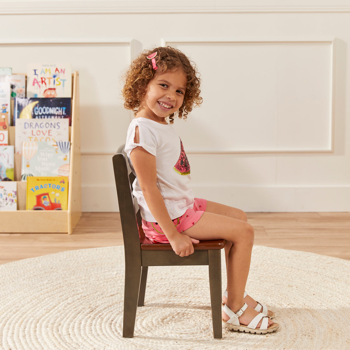 Child sitting on brown chair with red cushion in playroom setting with storage unit and books.