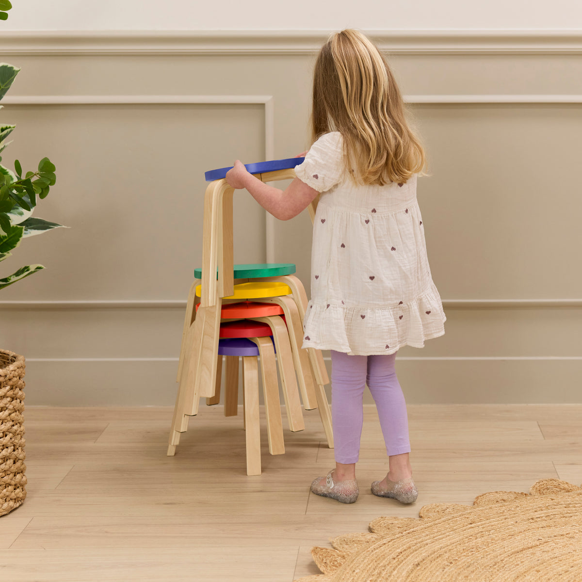 Child stacking six bentwood stools with colorful seats in blue, green, yellow, orange, red, and purple.